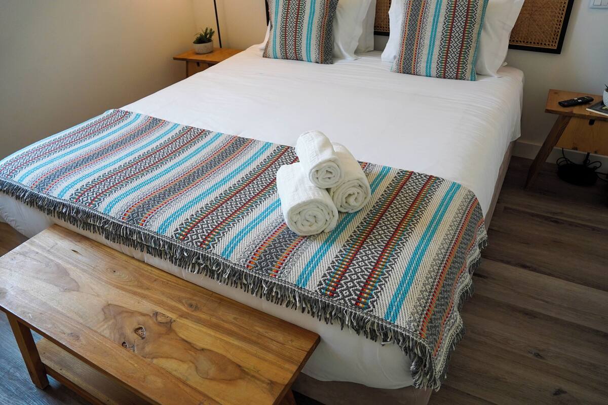 Ein ordentlich bezogenes Bett mit einer gemusterten Decke, zusammengerollten Handtüchern und einem Nachttisch aus Holz mit einer Fernbedienung.