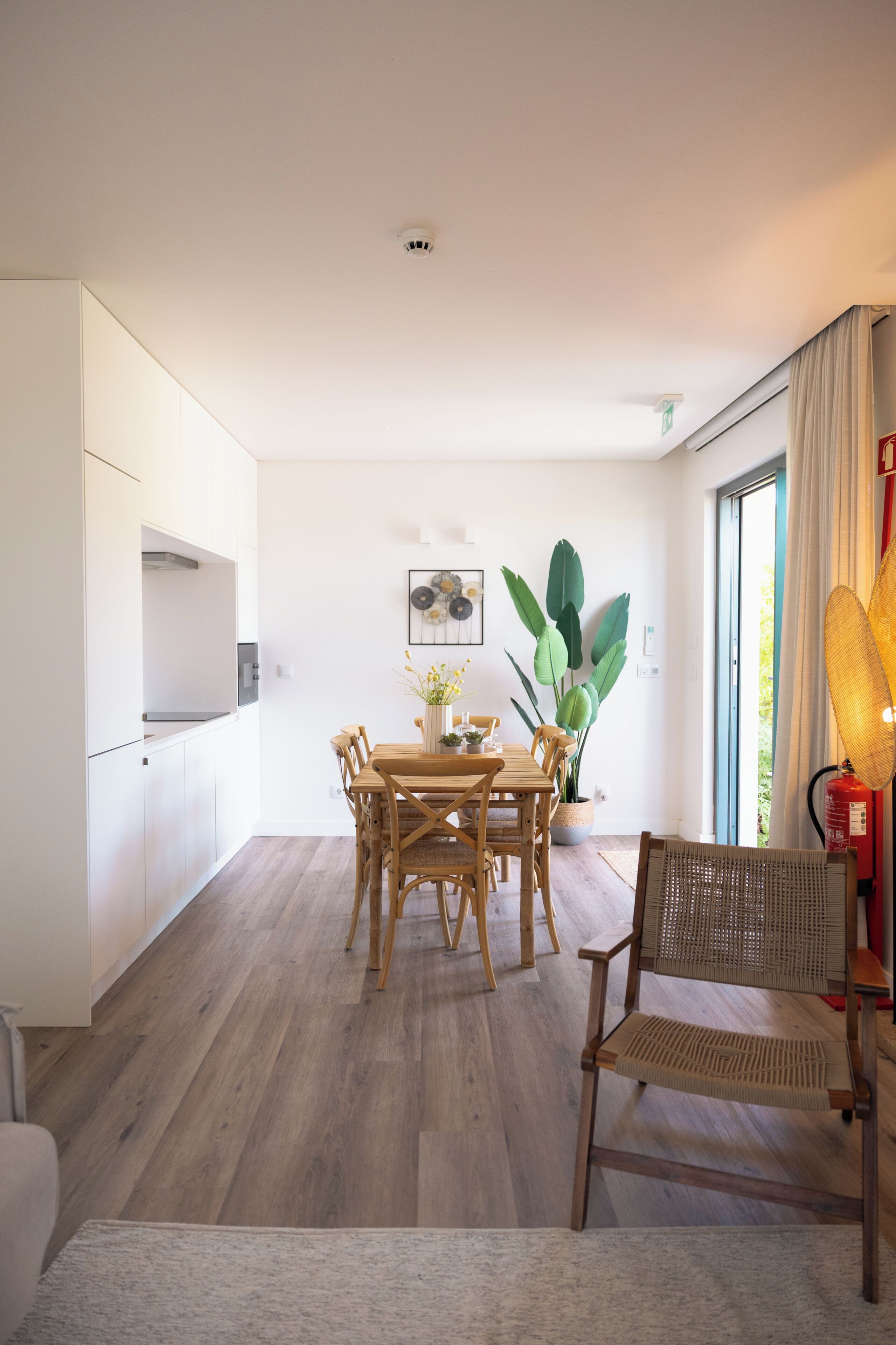 Eine moderne Küche mit weißen Schränken, einem Esstisch mit Stühlen, einem Blumentopf und einem großen Fenster mit Vorhängen.