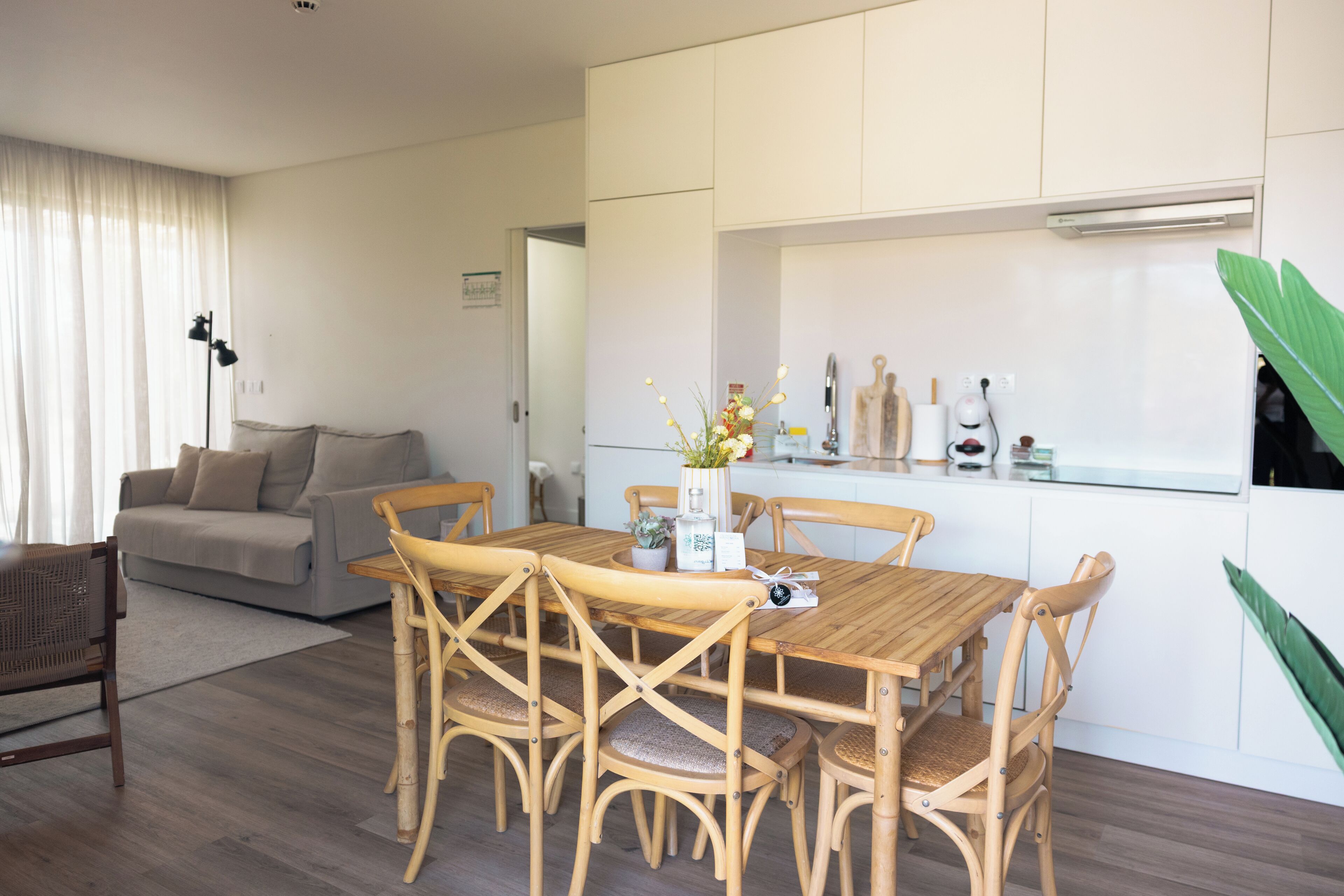 A dining area with a wooden table and chairs, a kitchen with white cabinets, and a living room with a sofa and a lamp.