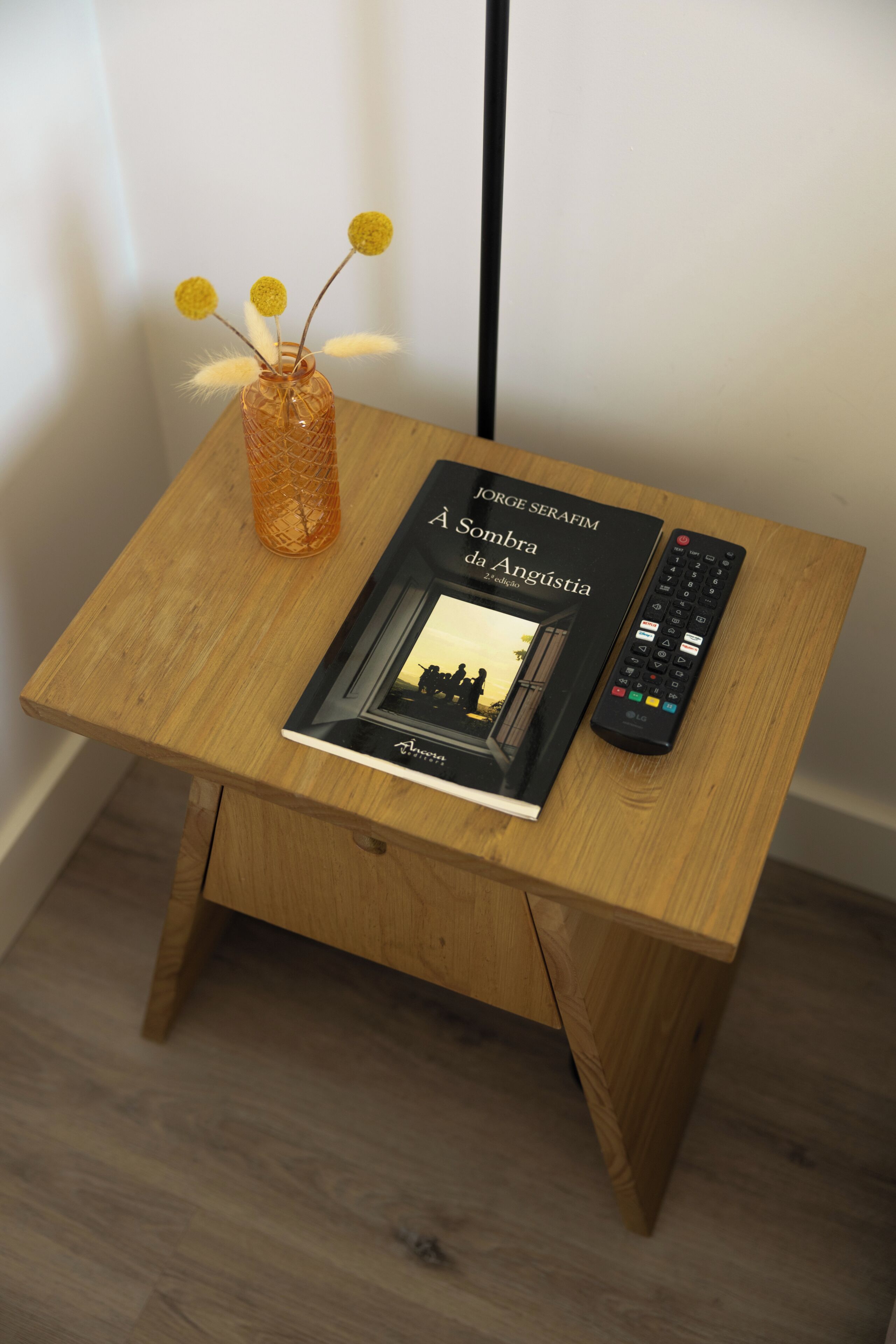 Un piccolo tavolo di legno con un libro intitolato 'A Sombra da Angústia' di Jorge Searafim e un telecomando.