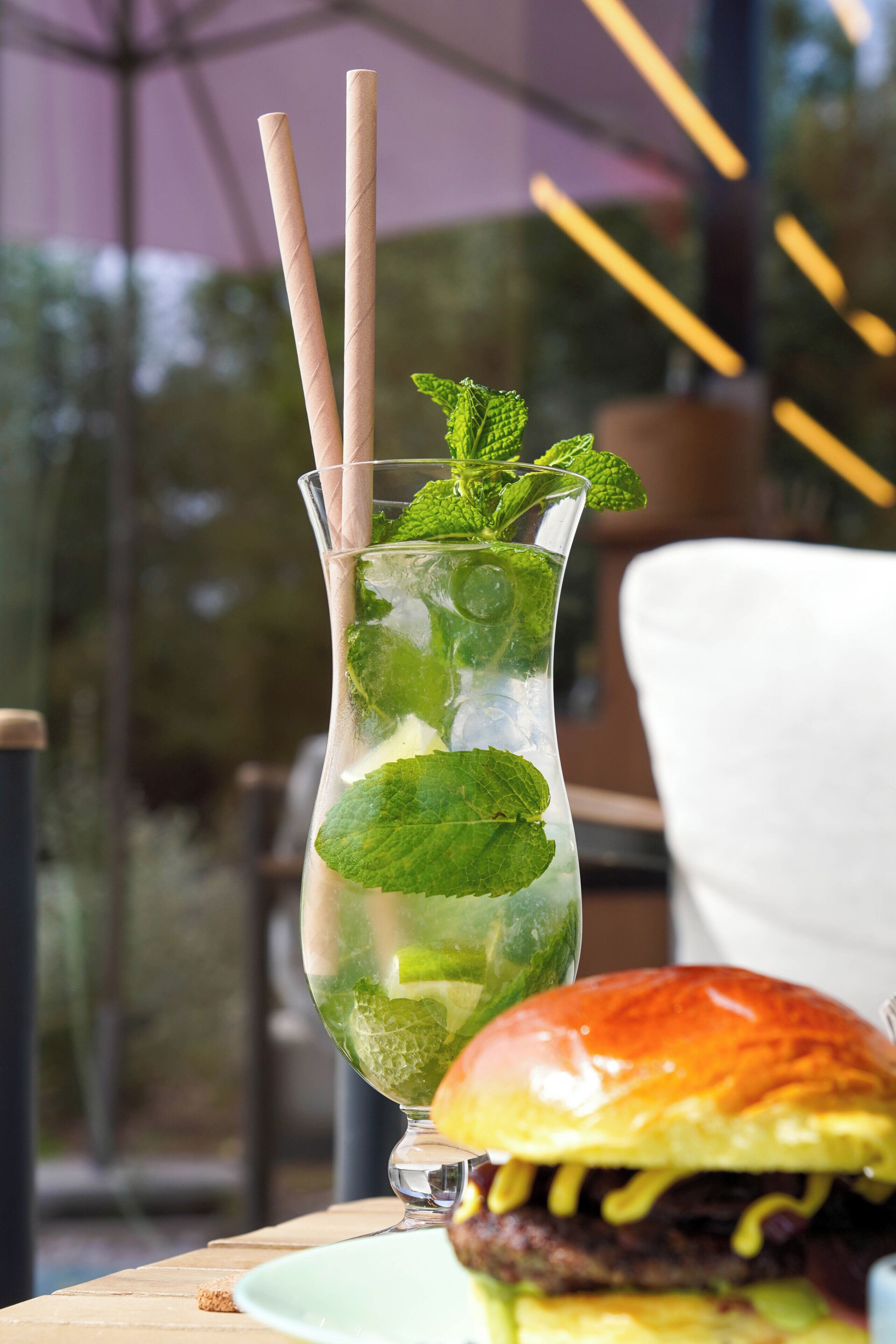 A glass with mint leaves and a straw, a burger, and a table in a restaurant setting.