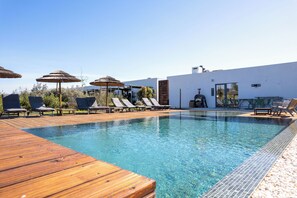 A clear swimming pool with lounge chairs and umbrellas, a wooden deck, and a white building with a door and windows.