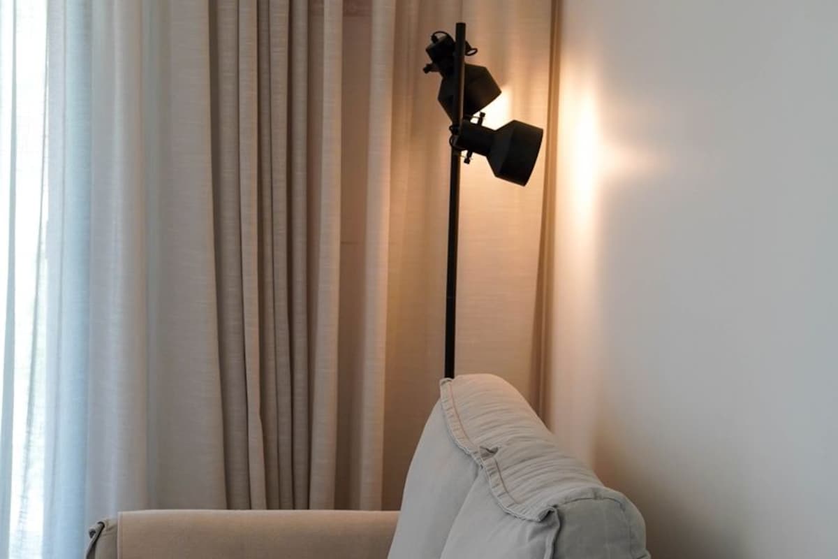 Un canapé aux couleurs neutres, agrémenté d’un plaid, d’une lampe sur pied et de rideaux légers, dans une pièce dont le mur est d’une couleur unie.