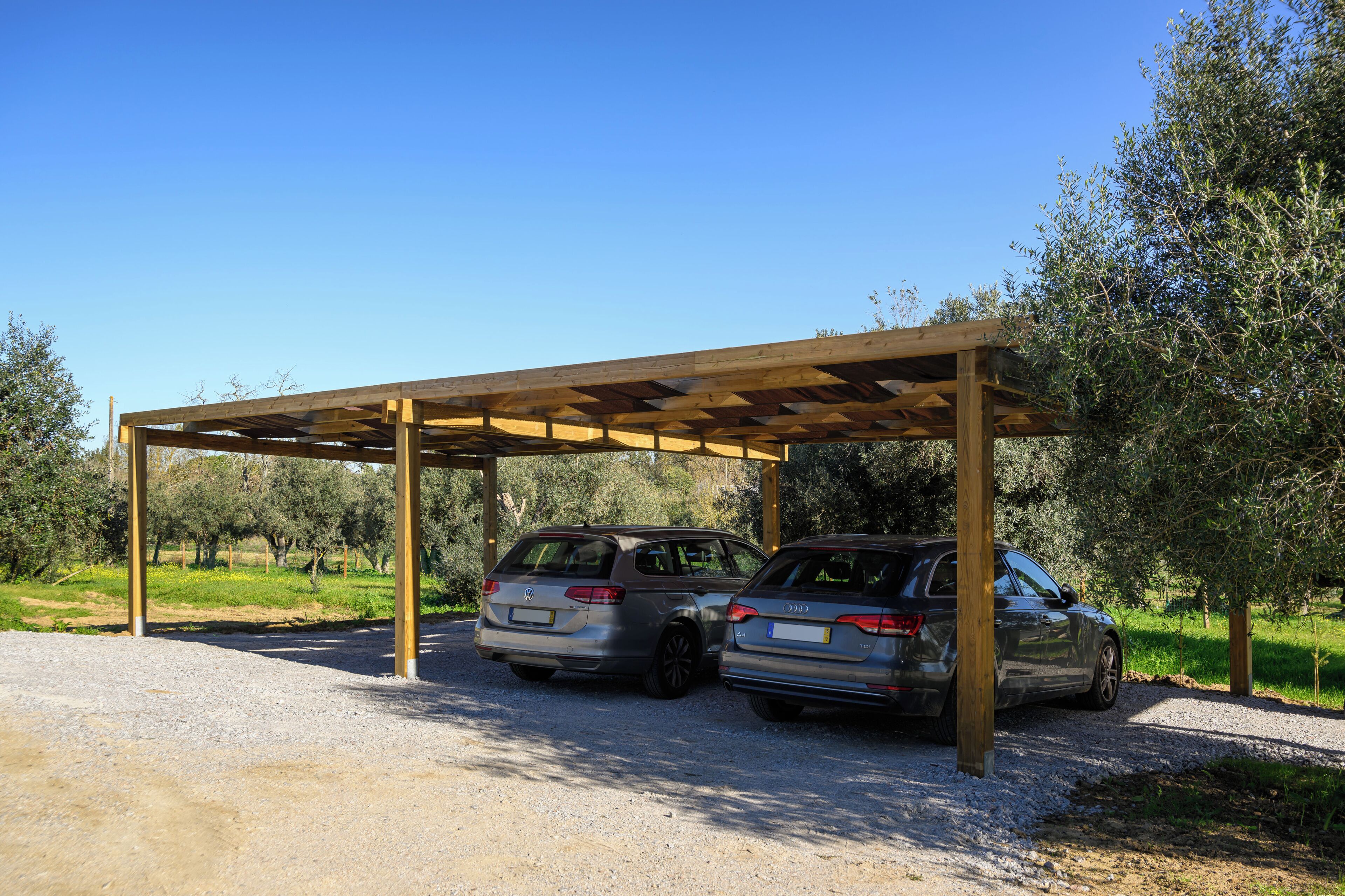Two cars parked under a wooden pergola in a rural setting with olive trees.