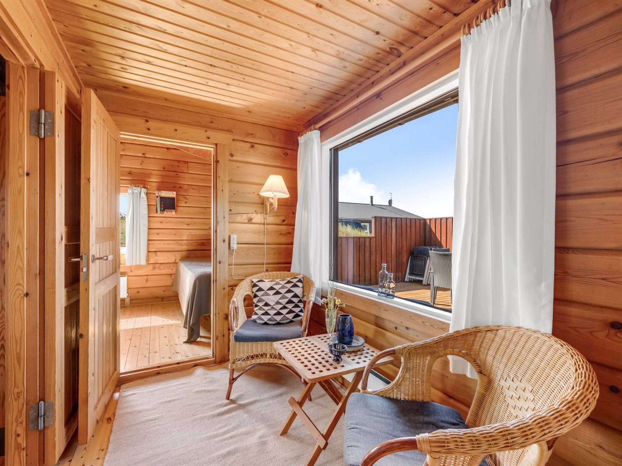 En stuga byggd av trä med en ombonad inredning, med ett matområde med möbler av rotting, ett fönster med gardiner och utsikt över omgivningen.