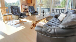A cozy living room with a wooden ceiling, large windows, a black leather sofa, two leather chairs, a wooden coffee table, and a small bookshelf.