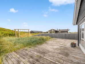Una terraza de madera que conduce a una casa moderna con un amplio ventanal, rodeada por una valla de madera y un área con césped.
