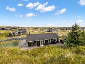 Una casa moderna con una amplia terraza, rodeada por un paisaje natural de dunas y vegetación escasa.
