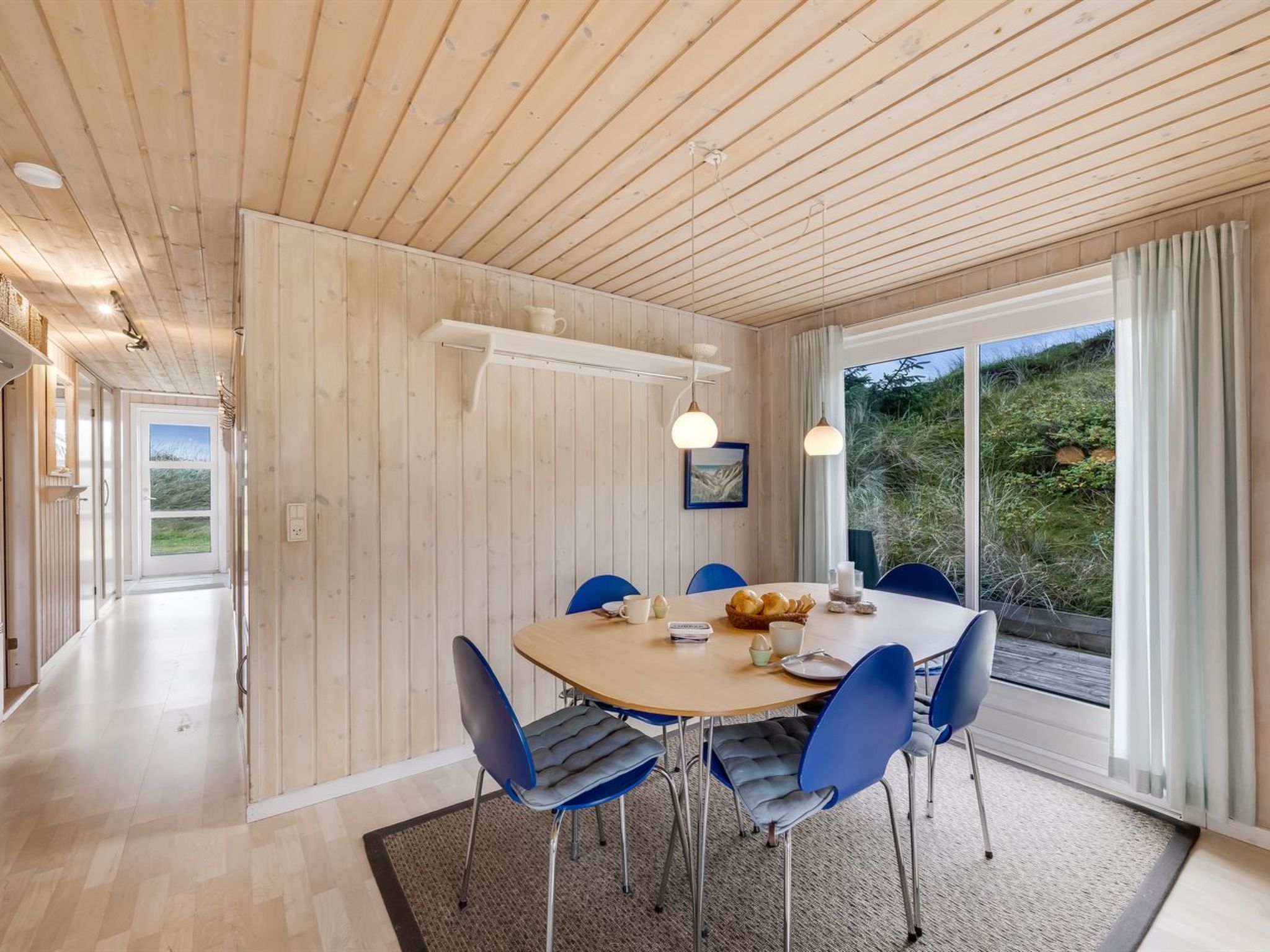 A modern dining area with a wooden ceiling, a large window with a view of greenery, and blue chairs around a wooden table.