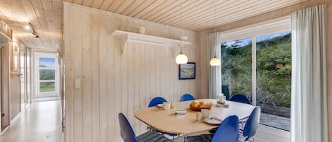 A modern dining area with a wooden ceiling, a large window with a view of greenery, and blue chairs around a wooden table.