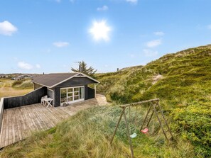 A wooden deck with a table and chairs, a small building with large windows, and a grassy hill in the background.