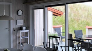 Une pièce avec un plafond en bois, une horloge, une étagère blanche remplie de livres, une table avec des chaises et une porte-fenêtre coulissante donnant sur un espace extérieur équipé de chaises et surmonté d’une structure à toit rouge.