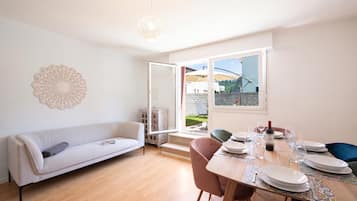 A modern living room with a sofa, dining table set for two, and a large window offering a view of the outdoors.