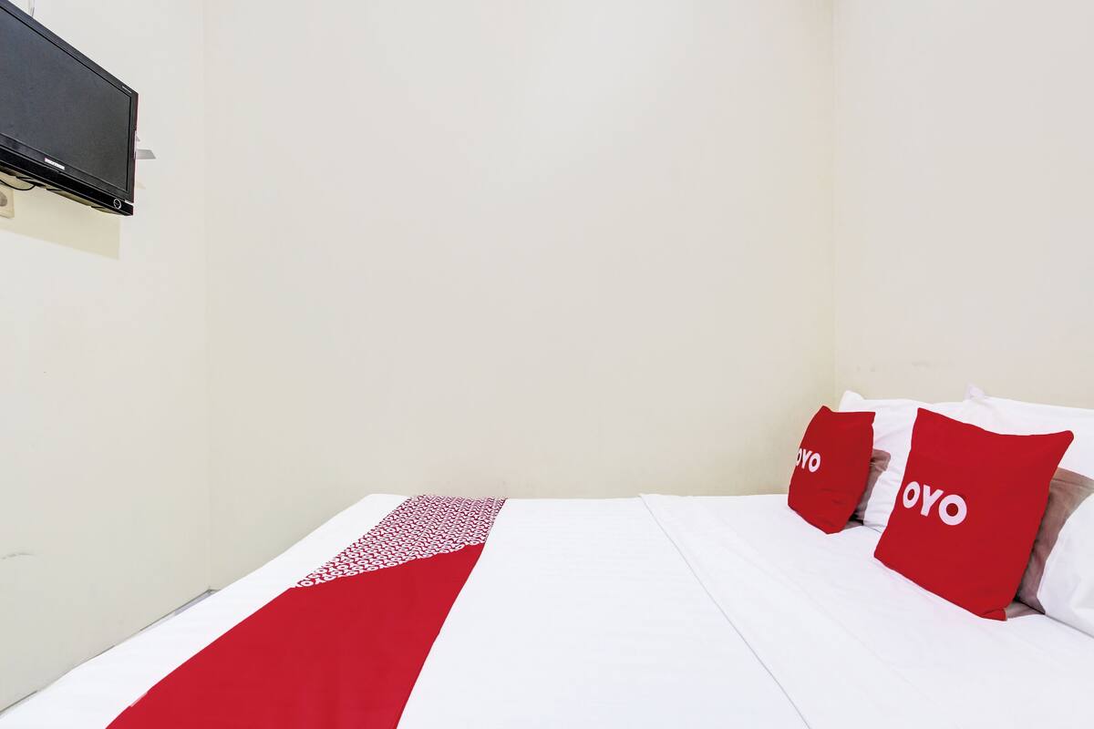 Ein Bett mit weißer Bettwäsche und roten Akzenten, ein an der Wand befestigter Flachbildfernseher und eine schlichte weiße Wand.