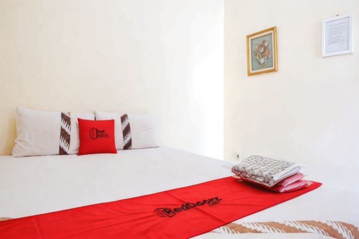 Una habitación de hotel con una cama, una manta roja y un cojín con un logotipo.