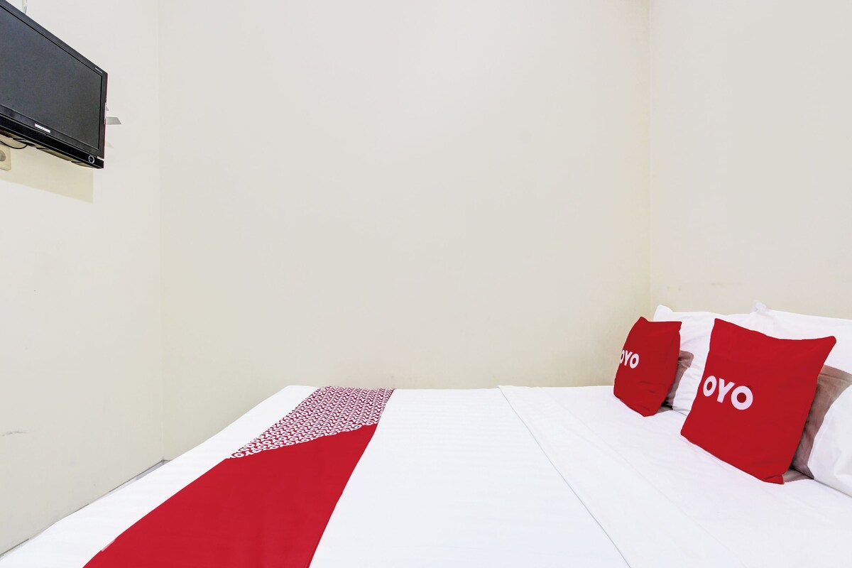 A bed with white bedding and red accents, a flat-screen TV mounted on the wall, and a plain white wall.