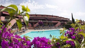 Un hotel con piscina rodeada de flores moradas, un edificio de varios pisos con balcones y un cielo despejado.