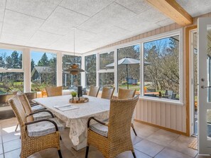Uma sala de sol com mesa de jantar, cadeiras de vime e grandes janelas com vista para o exterior.