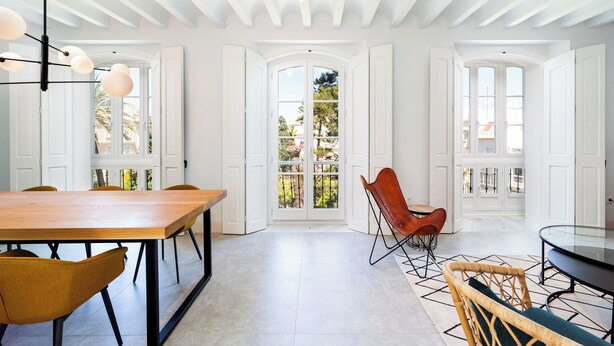 Un vaste espace à manger avec une longue table en bois et des chaises modernes, des portes blanches donnant sur un balcon et un plafond avec des poutres apparentes.