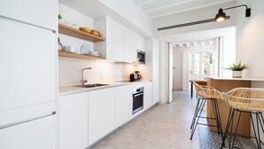 Une cuisine moderne avec des meubles blancs, un four encastré, un four à micro-ondes et une machine à café. On y trouve deux tabourets de bar en osier et une plante en pot sur le comptoir.