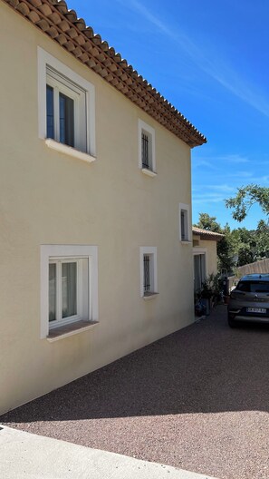 Ein zweistöckiges Gebäude mit einer beige farbenen Fassade und einem Ziegeldach. Drei Fenster mit weißen Rahmen und schwarzen Sprossen sind vorhanden. Ein Auto steht auf der Auffahrt.