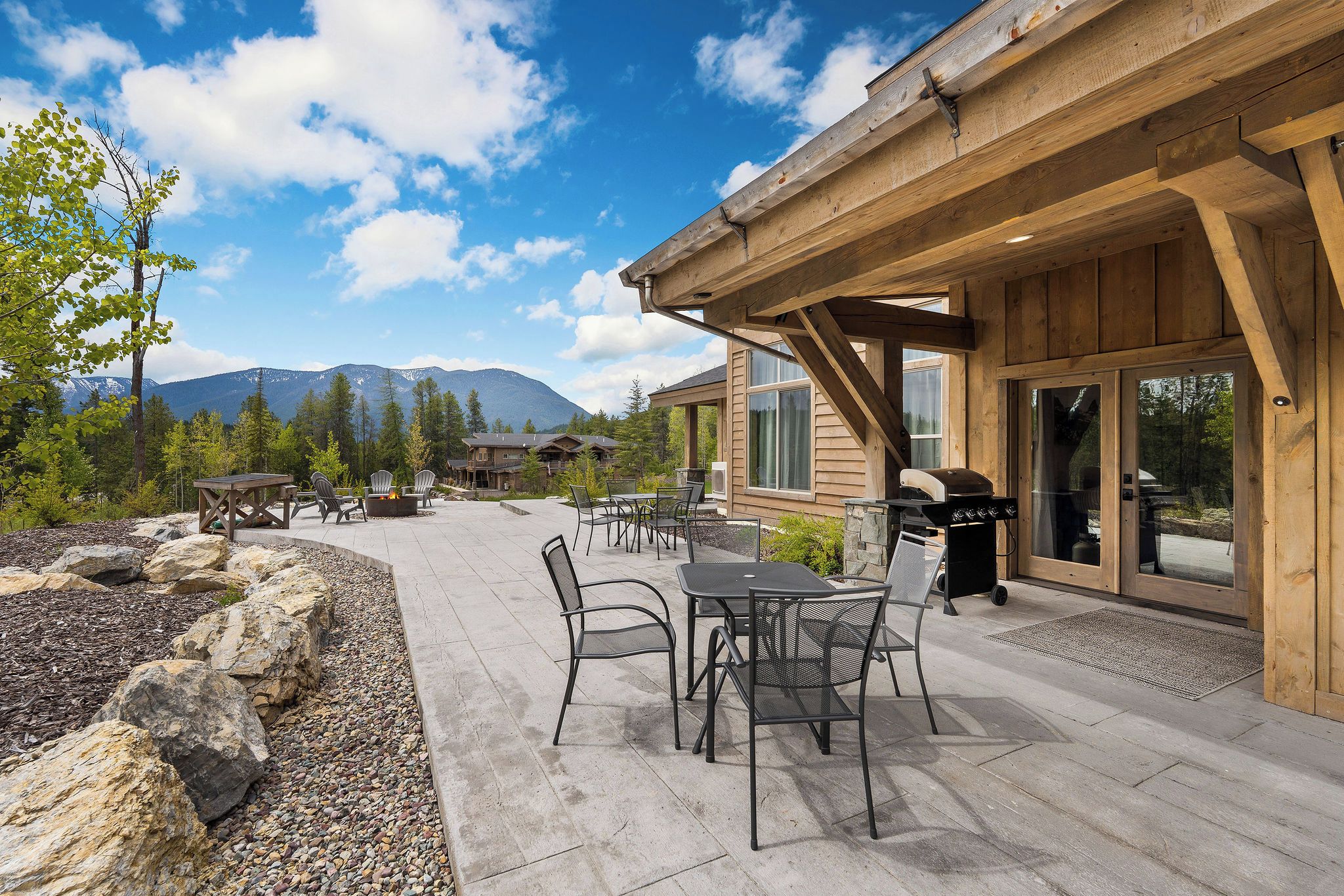 An outdoor patio with stone seating and a grill area, surrounded by a wooden deck and a view of mountains.