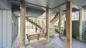 Un espace extérieur abrité, doté d’un escalier en bois menant à une terrasse, d’un sol en béton et d’une balançoire.