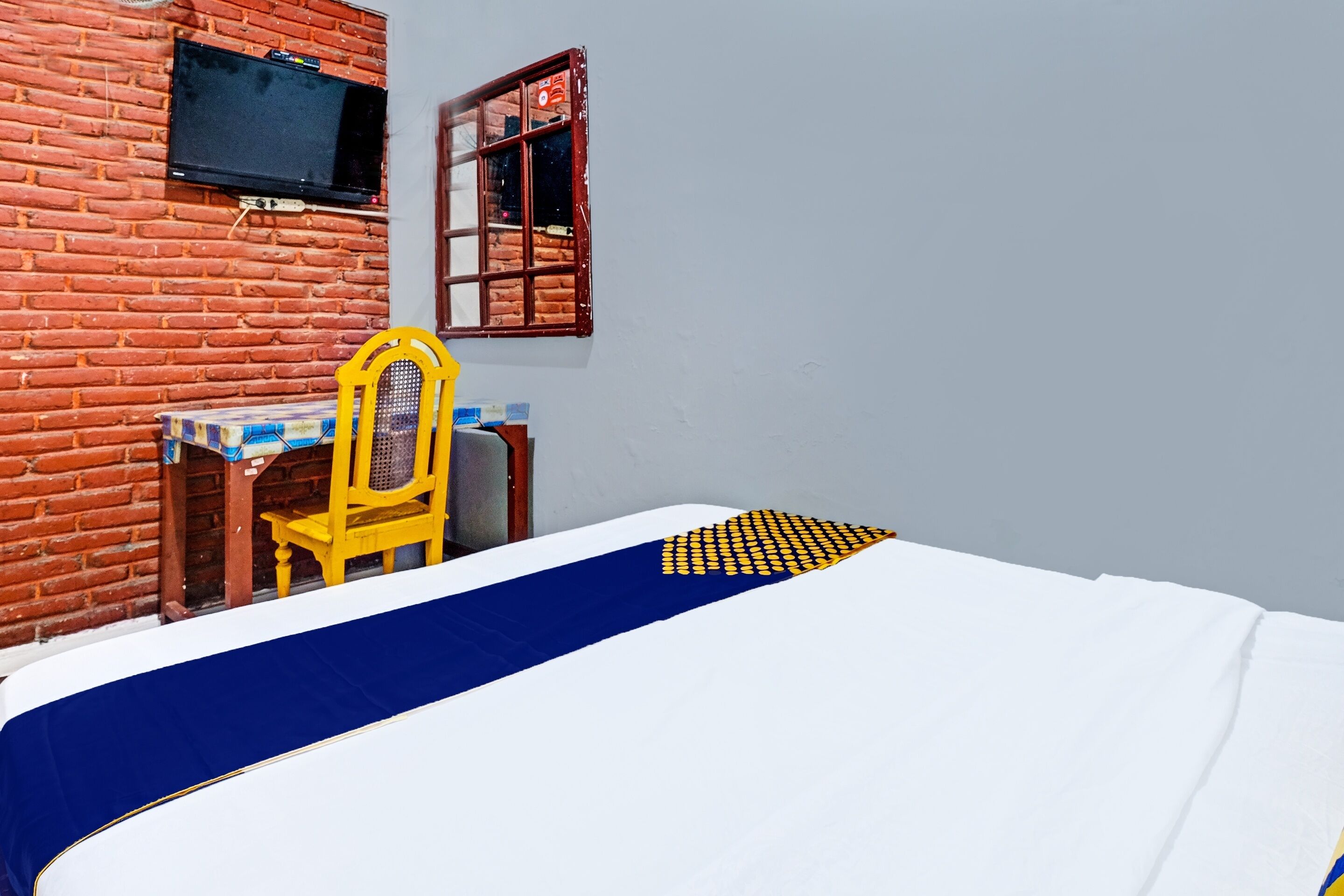 Une pièce comprenant un lit, un bureau, une chaise, un téléviseur fixé au mur en briques et une fenêtre avec des volets en bois.