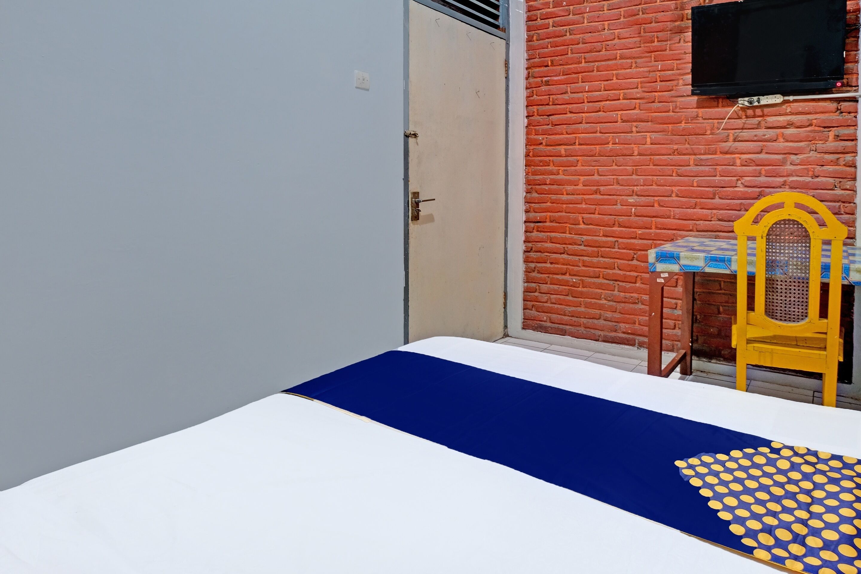A room with a bed, a chair, a small table, and a TV mounted on a brick wall.