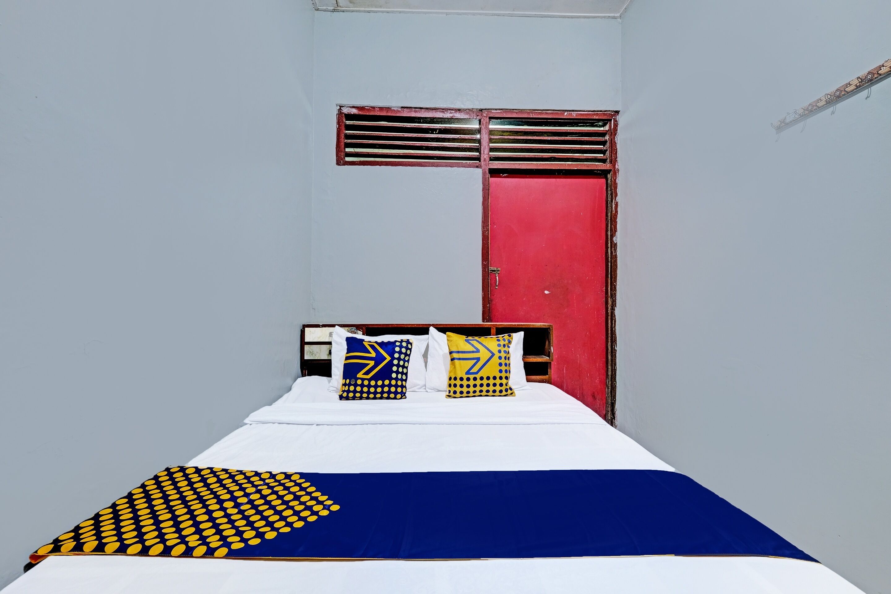A single bed with a white and blue bedspread, two pillows with geometric patterns, and a red door with a window above it.