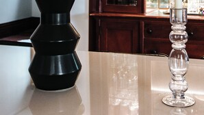A black vase with green foliage on a reflective surface, a clear glass candle holder with a white candle, and a wooden cabinet with glassware in the background. - LUX Uptown Escape - The Trester
 (Sheboygan)