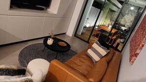 A modern living room with a brown leather sofa, a white armchair, and a black coffee table. There is a flat-screen TV mounted on the wall and a large window providing a view of the outdoors.