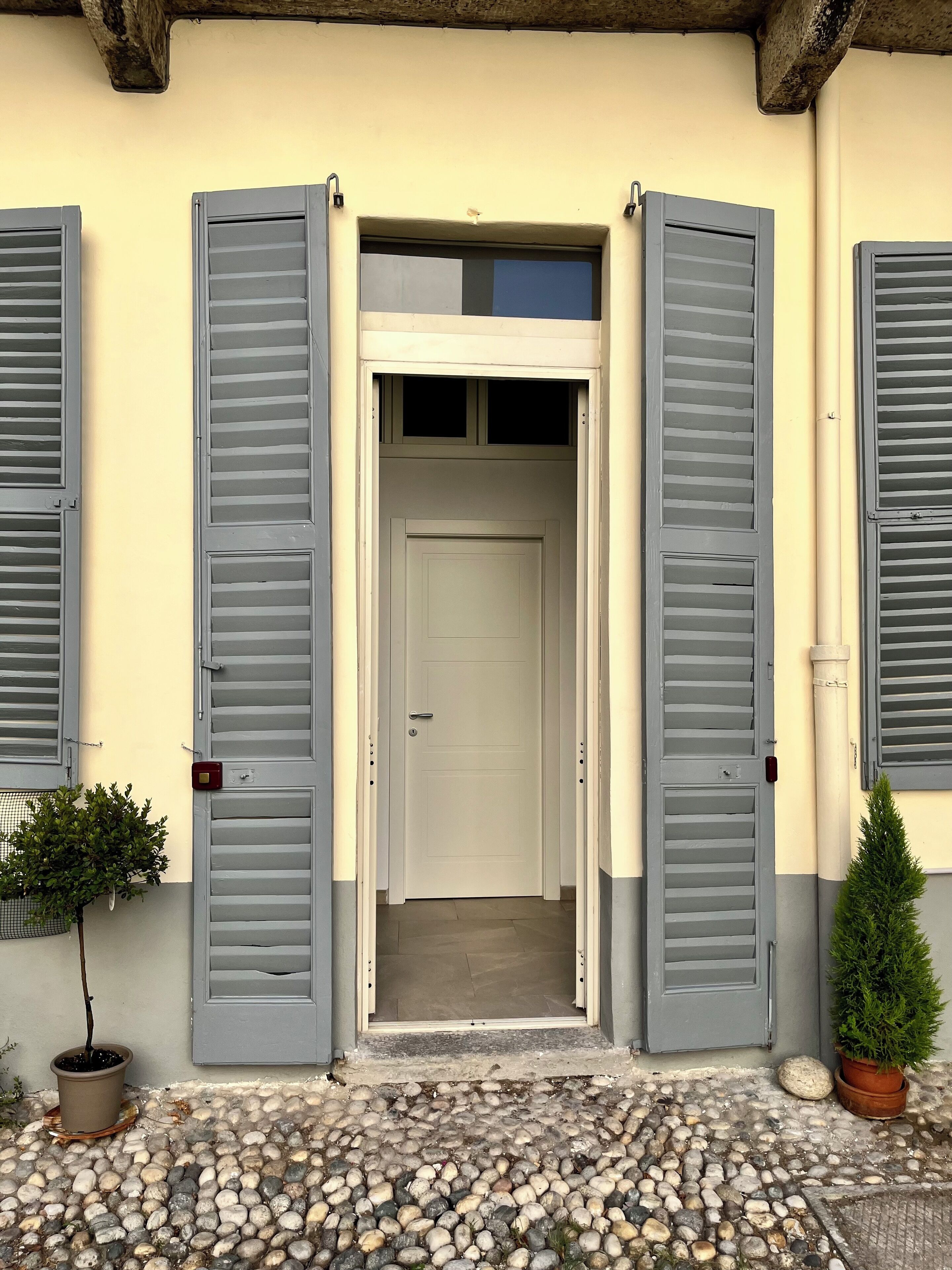 Ein Gebäudeeingang mit einer weißen Tür, grauen Fensterläden und einem gepflasterten Vorplatz.