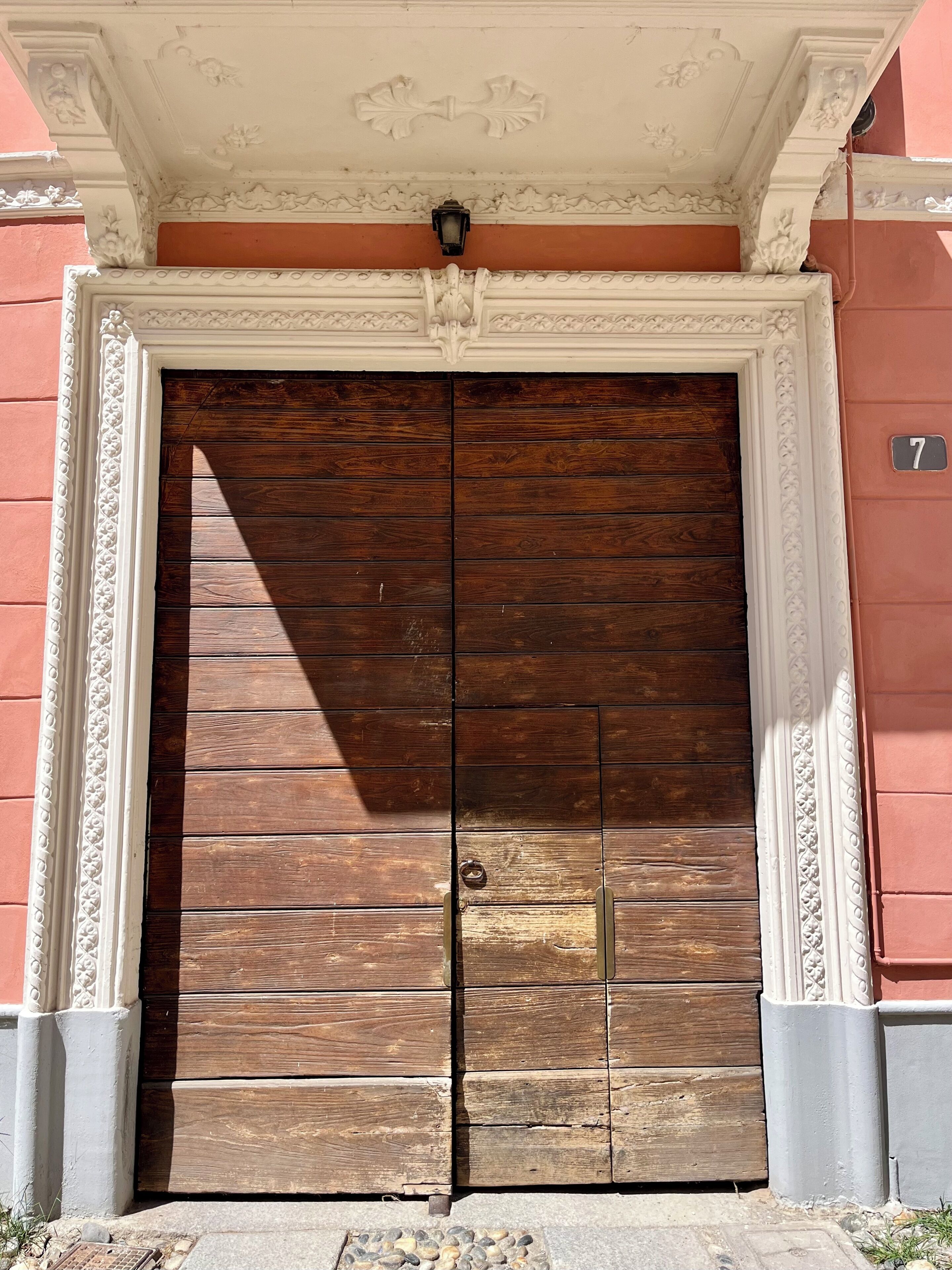 Una puerta doble de madera con un picaporte de latón, enmarcada por un elaborado moldura blanca, sobre un pared rosa con un letrero con el número '7'.