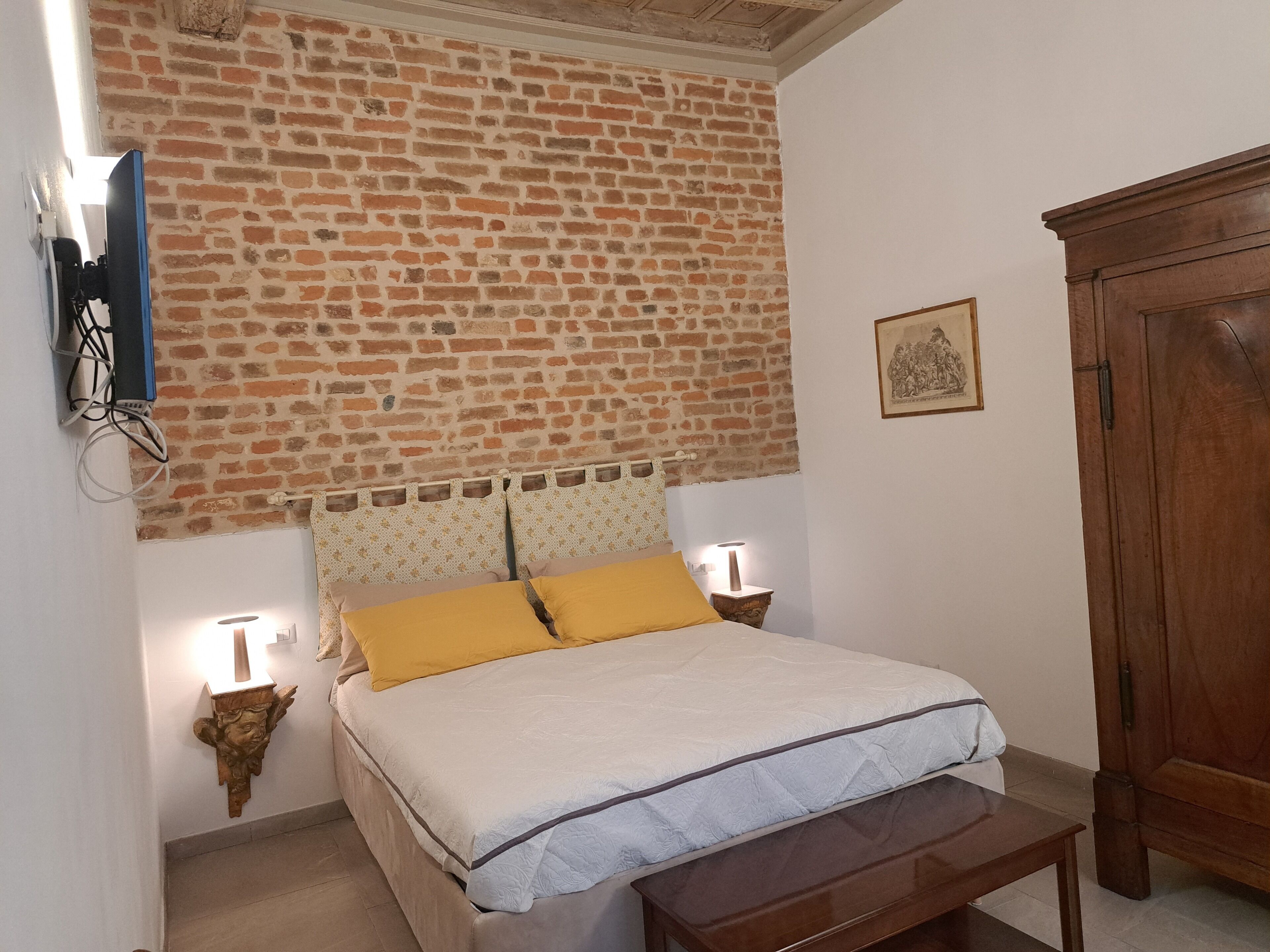 A bedroom with a brick wall, a bed with a white and blue bedspread, two bedside lamps, a wooden cabinet, and a wall-mounted TV.