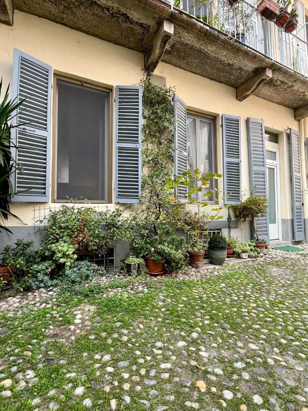 A courtyard with a paved ground, potted plants, and a building with multiple windows and shutters.