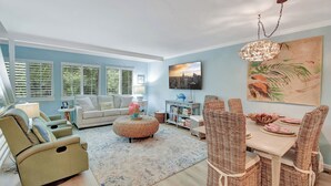 Un salon comprenant un canapé, un fauteuil, un pouf, une table à manger avec des chaises, un téléviseur et des œuvres d’art accrochées au mur.