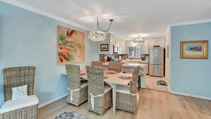 Un coin repas avec des chaises en osier et une table, une cuisine avec des meubles blancs et un plafond agrémenté d’un lustre.