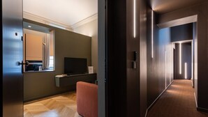 Ein modernes Hotelzimmer mit Flachbildfernseher, einem kleinen Tisch und einer Couch. Das Zimmer verfügt über einen Holzboden im Fischgrätmuster und einen großen Spiegel.