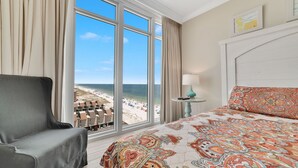 Camera d'albergo con un'ampia finestra vista spiaggia, un letto con copriletto decorato, una poltrona grigia e un tavolino con una lampada.