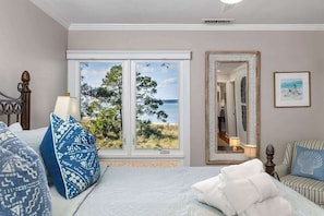 A bedroom with a large window, a bed, a chair, a mirror, and a framed picture on the wall.