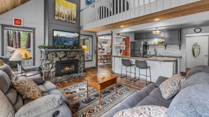 A spacious living area with a stone fireplace, a large sofa, and a dining area with a kitchen in the background. - New! Hot Tub ~ Heart of Sunriver ~  Fireplace ~ Updated Home ~ SHARC passes! (Sunriver)