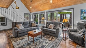 A spacious living room with a high wooden ceiling, grey sofas, a patterned rug, and large windows offering a view of the outdoors. - New! Hot Tub ~ Heart of Sunriver ~  Fireplace ~ Updated Home ~ SHARC passes! (Sunriver)