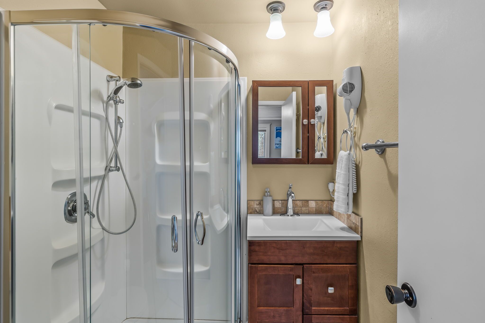 Une salle de bain dotée d’une douche à parois de verre, d’un meuble-lavabo avec un évier, d’un miroir et d’un sèche-cheveux fixé au mur.