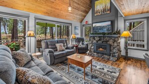A cozy living room with a stone fireplace, grey sofas, and a large window overlooking a forest. - New! Hot Tub ~ Heart of Sunriver ~  Fireplace ~ Updated Home ~ SHARC passes! (Sunriver)