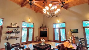 Uma sala de estar ampla com teto de madeira alto, um candelabro, uma lareira e grandes janelas.