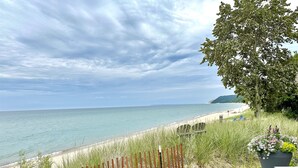 Une terrasse en bois surplombe une plage dont le rivage est sablonneux, et une zone herbeuse est délimitée par une clôture en bois.