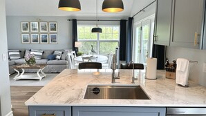 Une cuisine moderne dotée d’un plan de travail en marbre, d’un évier en acier inoxydable et de luminaires suspendus. On y trouve un coin repas avec une table et des chaises blanches, ainsi qu’un salon avec un canapé et des coussins décoratifs.