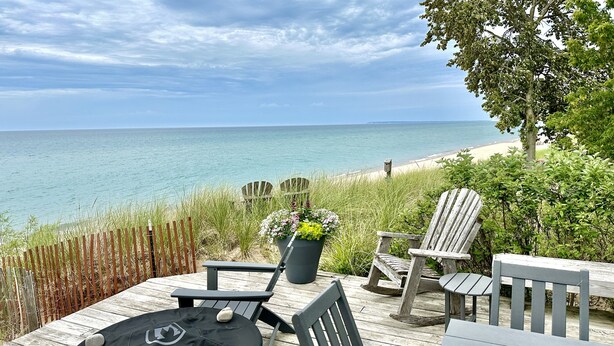 Une terrasse en bois avec des fauteuils Adirondack, une table et un pot de fleurs, le tout offrant une vue sur une plage et l’océan.