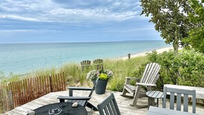 A wooden deck with Adirondack chairs, a table, and a potted plant overlooking a beach and ocean. - Empire Beach House - Lake Michigan cottage with firepit, grill & furnished deck (Empire)